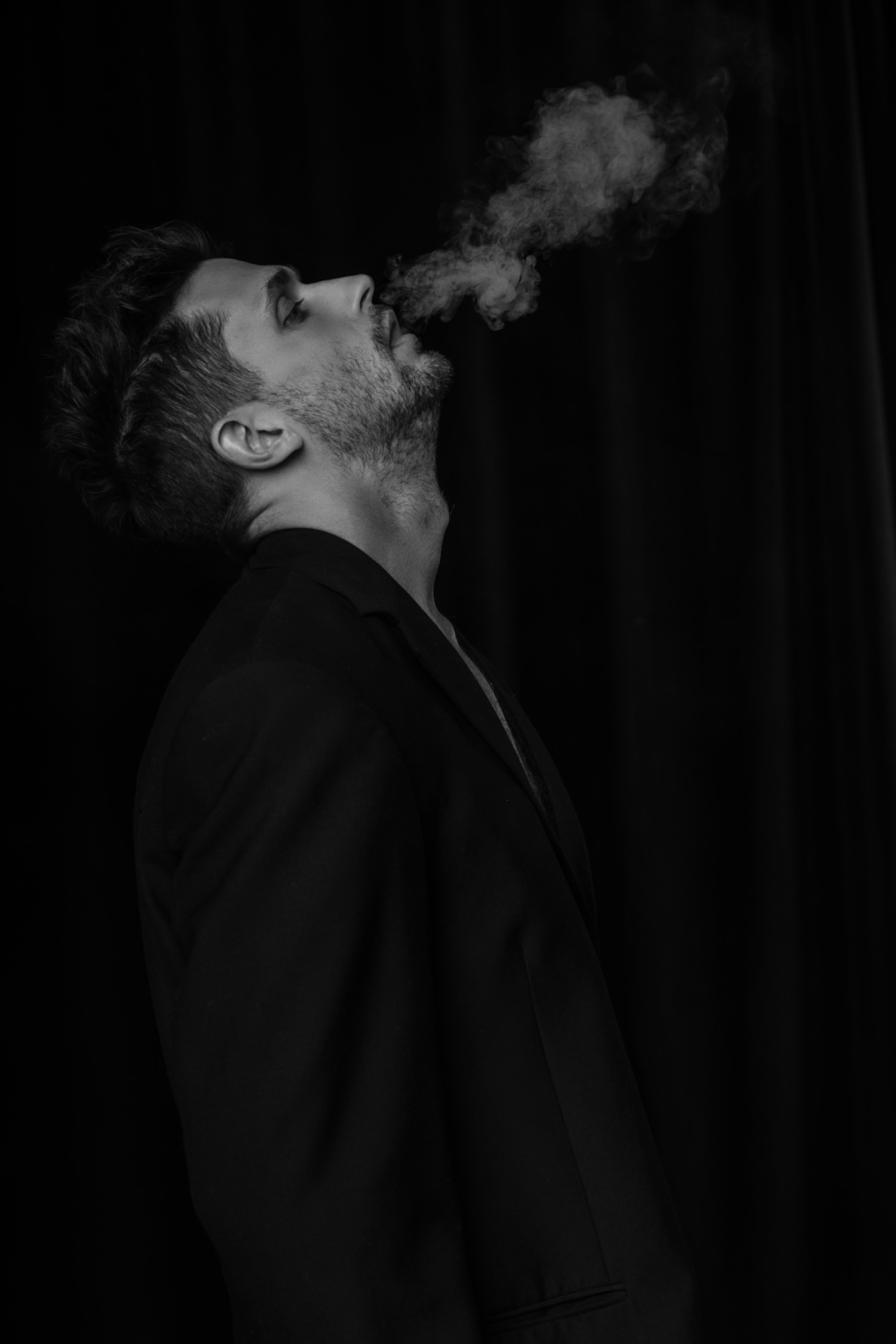 Czarno-białe zdjęcie profilowe mężczyzny w marynarce, z głową odchyloną do tyłu, wydmuchującego dym w górę na czarnym tle.