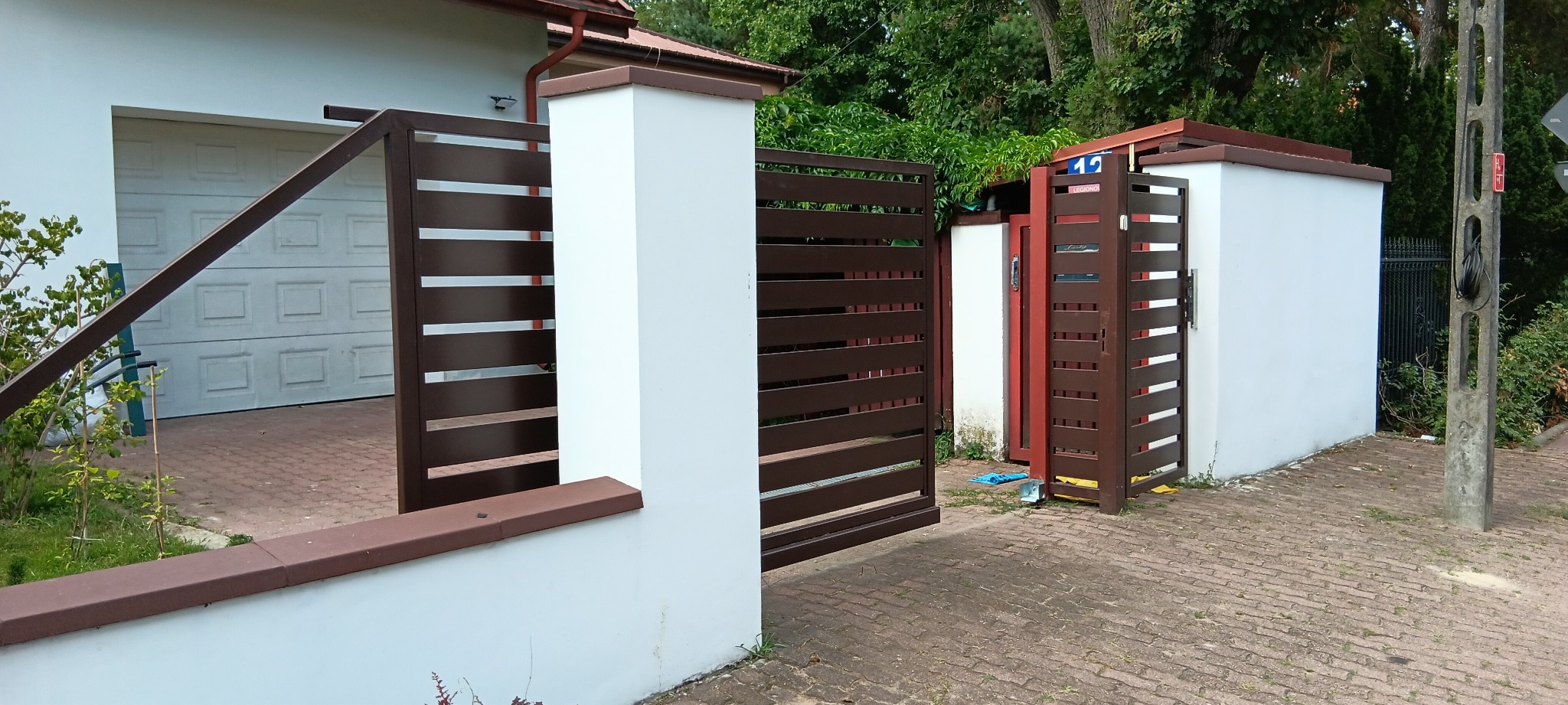 Nowoczesne, brązowe, poziome panele ogrodzeniowe i brama wjazdowa z białymi słupkami, widoczna brama garażowa w tle.