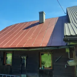 Czerwony dach z blachy na drewnianym domu z kominem, widoczne okna i elementy elewacji, błękitne niebo w tle.