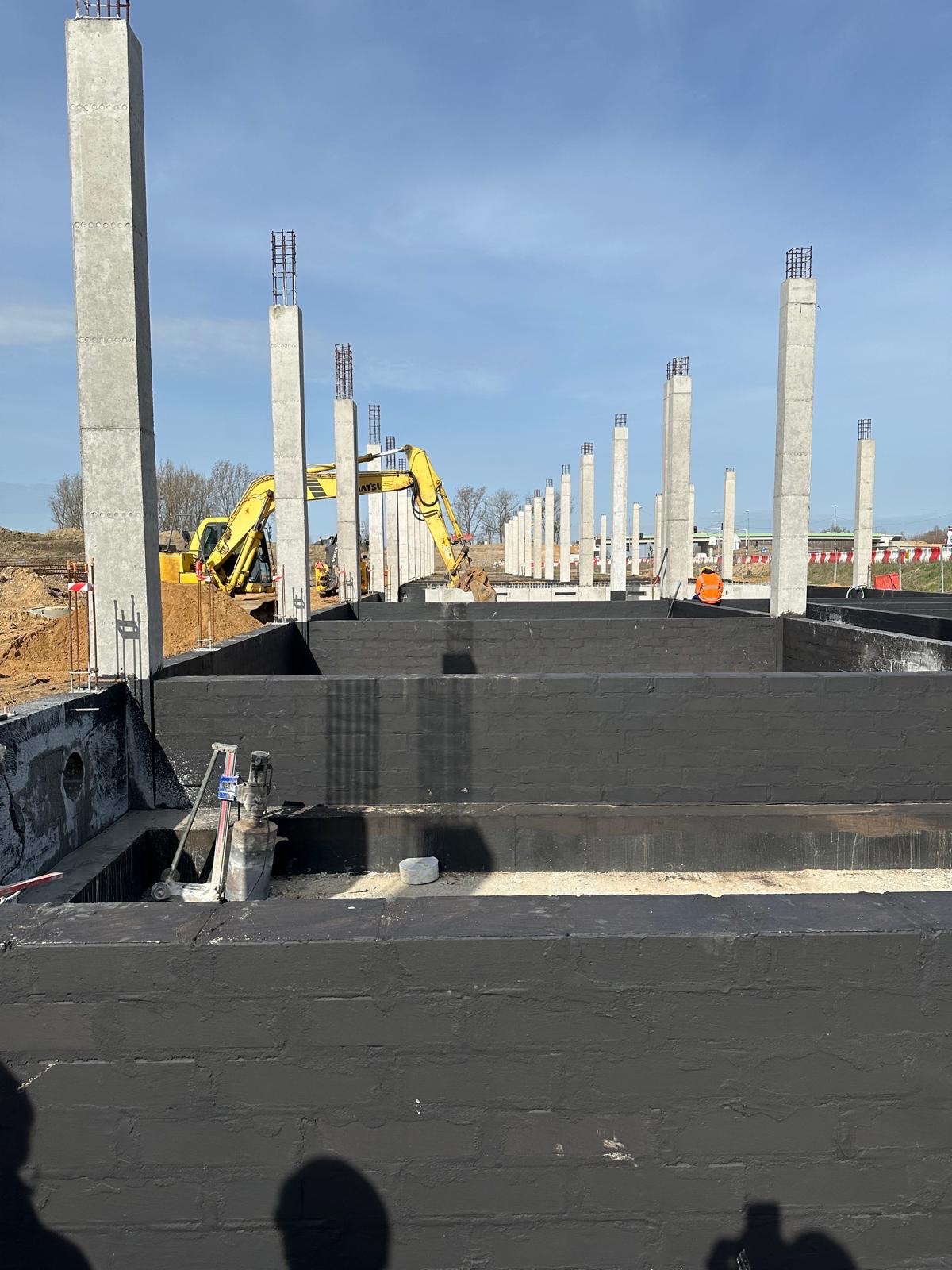 Wykopane fundamenty z izolacją bitumiczną, widoczne betonowe słupy z wystającymi prętami zbrojeniowymi oraz koparka w tle.