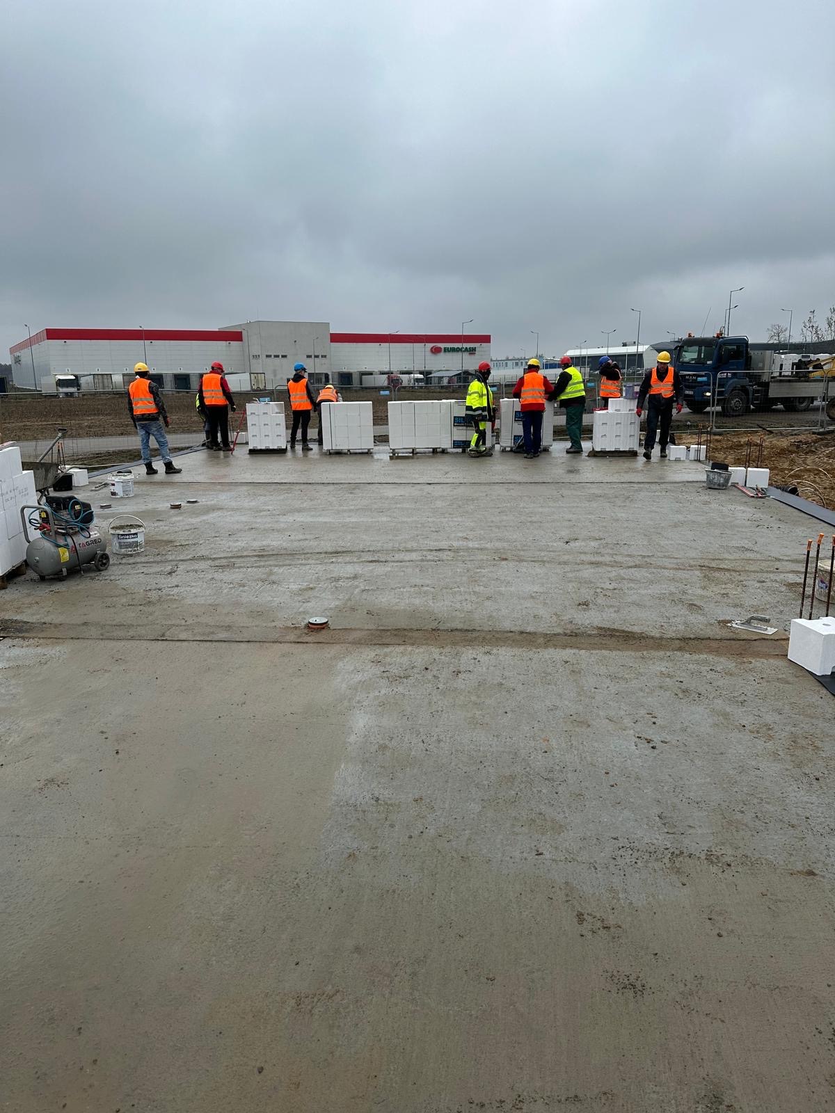 Przygotowanie fundamentów budynku usługowego: grupa robotników w odblaskowych kamizelkach ustawia bloczki Ytong na wylanej płycie betonowej, w tle widoczny budynek Eurocash i sprzęt budowlany...
