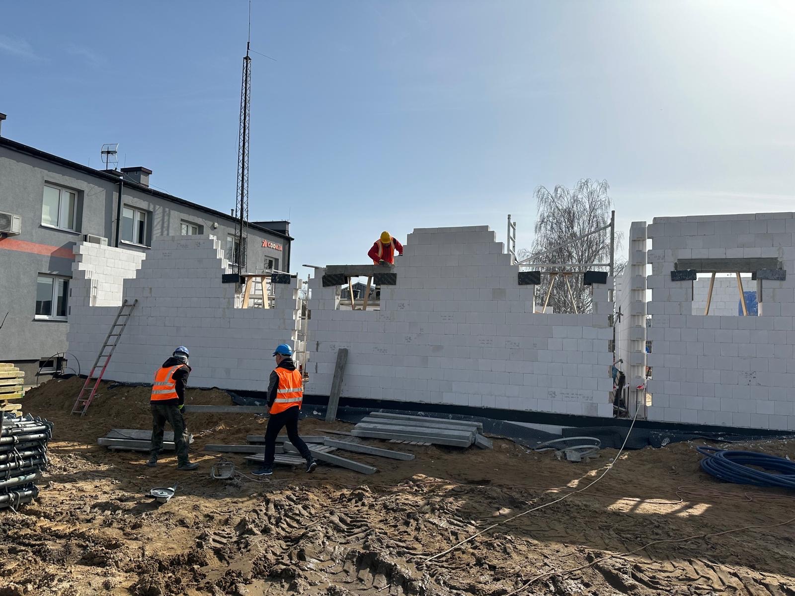 Budowa domu z białych bloczków: murarze w pomarańczowych kamizelkach pracują na placu budowy, w tle budynek z anteną i drzewo bez liści.