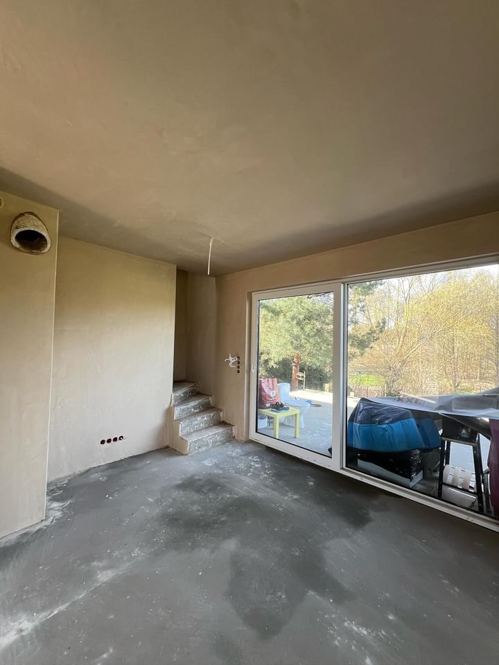 Wnętrze w trakcie prac wykończeniowych, ściany pokryte świeżym tynkiem, widoczne schody w surowym stanie i duże okno przesuwne z widokiem na zewnątrz.