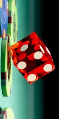 Przezroczysta czerwona kostka do gry z białymi oczkami, ustawiona pod kątem, oparta o zielone i niebieskie żetony do gry na tle czerni.