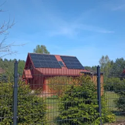 Czerwony dom z panelami słonecznymi na dachu, widoczny zza czarnego metalowego ogrodzenia, z drzewami w tle i błękitnym niebem.