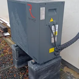 Szara pompa ciepła marki Rotenso umieszczona na betonowych blokach, widoczne przyłącza, otoczenie posesji z kamiennym podłożem i fragmentem elewacji budynku.