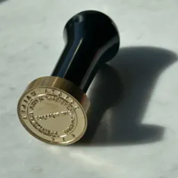 Złoty stempel z czarną rączką na marmurowym tle, rzucający wyraźny cień.