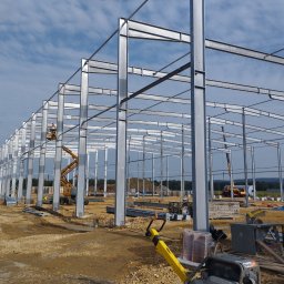 RAK BUD PIOTR RAK - Stalowa konstrukcja hali w trakcie montażu, widoczny dźwig koszowy i pracownicy w oddali, teren budowy z widocznym kominem w tle.