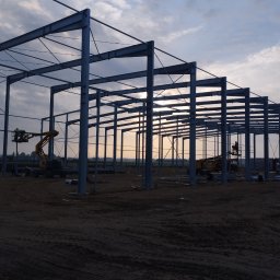 RAK BUD PIOTR RAK - Konstrukcja stalowa hali w trakcie budowy, widoczne słupy i belki, dwie maszyny budowlane z pracownikami na podnośnikach, ziemisty teren budowy, zachmurzone niebo w tle.