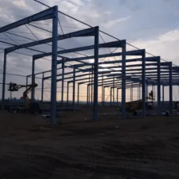 Konstrukcja stalowa hali w trakcie budowy, widoczne słupy i belki, dwie maszyny budowlane z pracownikami na podnośnikach, ziemisty teren budowy, zachmurzone niebo w tle.