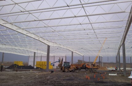 Konstrukcja stalowa hali w trakcie budowy, widoczne dźwigi, koparka i materiały budowlane na placu budowy w pochmurny dzień.