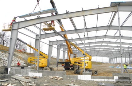 Montaż konstrukcji stalowej hali z użyciem podnośników koszowych, widoczne płyty warstwowe w trakcie instalacji na szkielet konstrukcji.