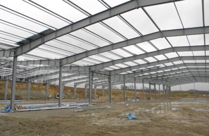 Konstrukcja stalowa hali w trakcie budowy w Nowej Wsi, widoczne słupy i elementy dachu, na ziemi leżą materiały budowlane i stoi koparka.