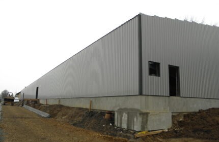 Długa hala przemysłowa z szarej blachy trapezowej, widoczna w trakcie budowy, z fundamentem betonowym i otworami na okna i drzwi. Na pierwszym planie ziemia i mała koparka.