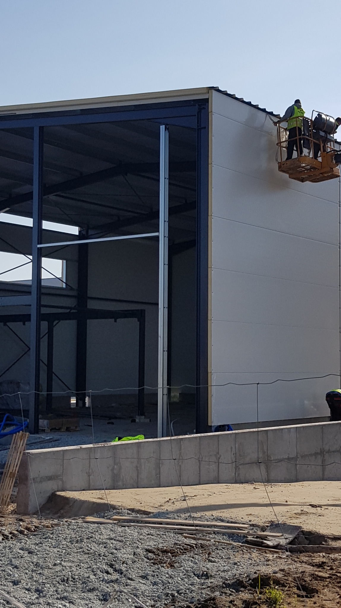 Wznoszona hala z szarej płyty warstwowej z widoczną stalową konstrukcją, pracownicy na podnośniku koszowym montują elementy na wysokości.