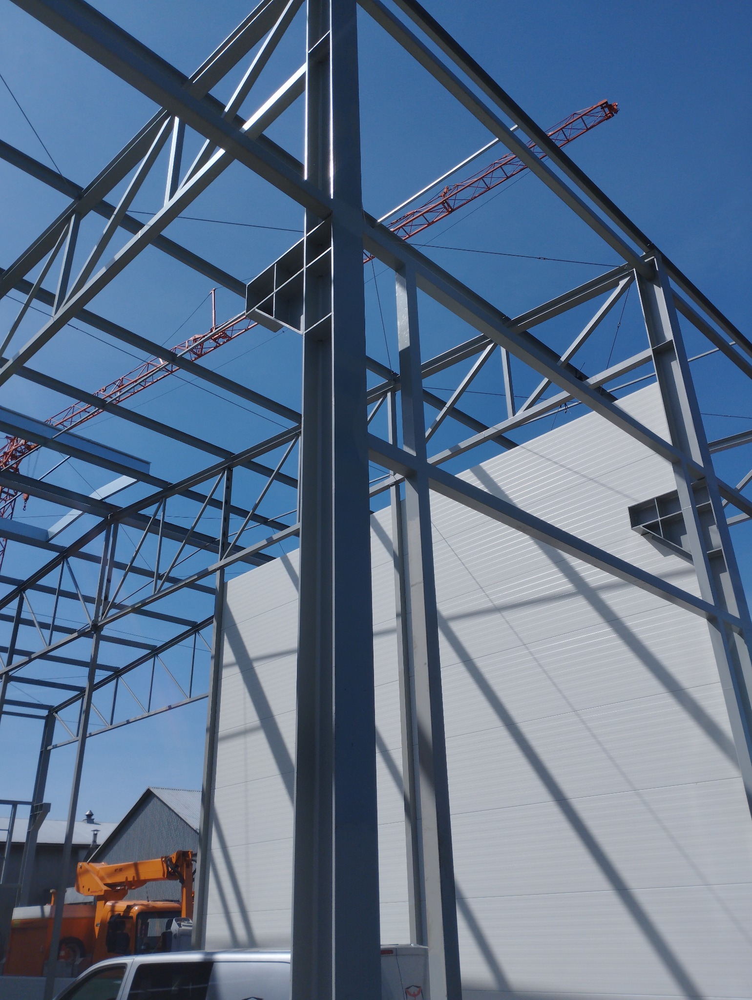 Szara konstrukcja stalowa hali w trakcie budowy, widoczny żuraw na tle błękitnego nieba i fragment ściany z płyty warstwowej.
