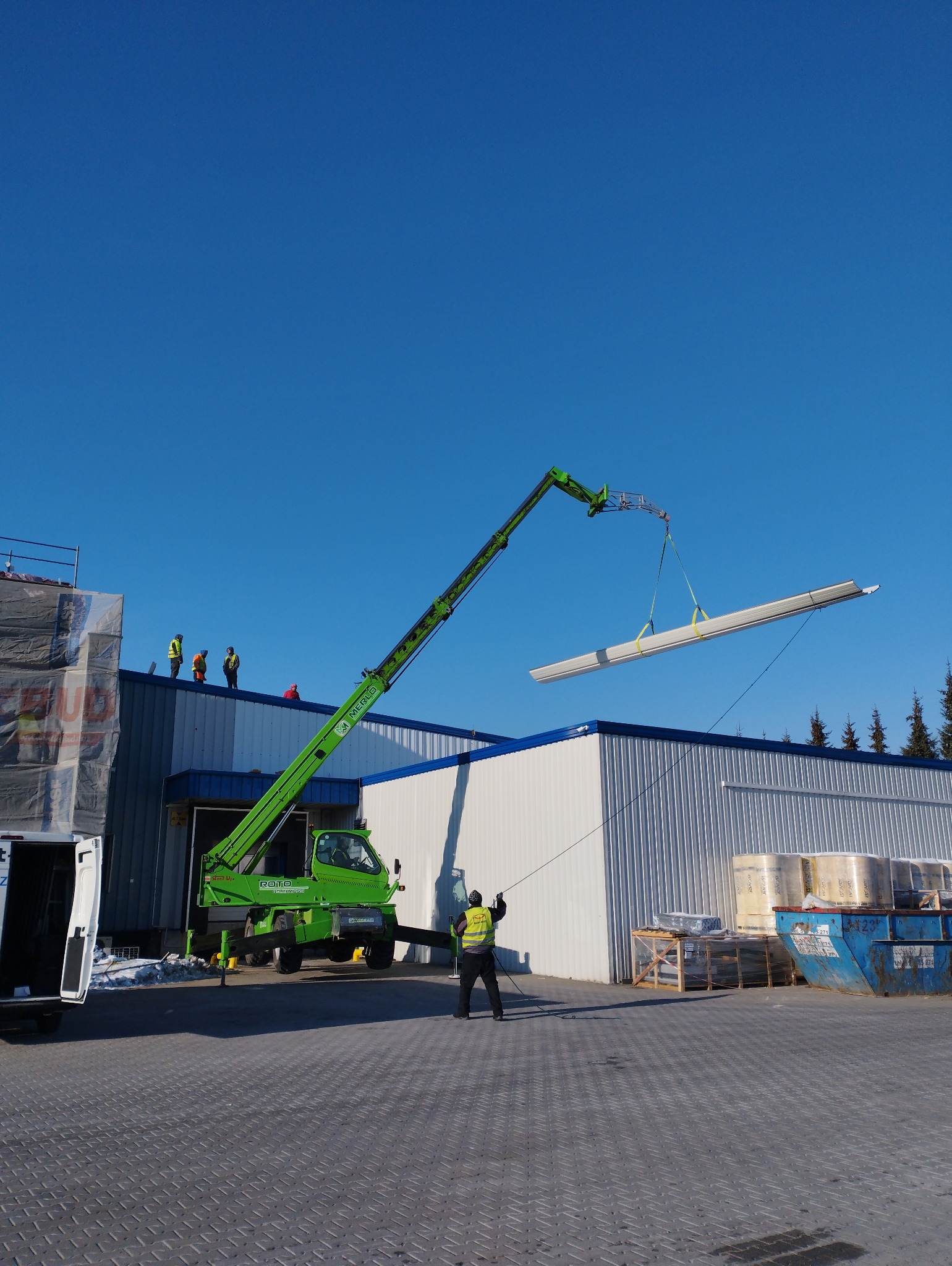 Zielony dźwig teleskopowy podnosi pakiet płyt na tle hali z blachy trapezowej, pracownik w kamizelce odblaskowej nadzoruje operację, na dachu widoczni inni pracownicy.