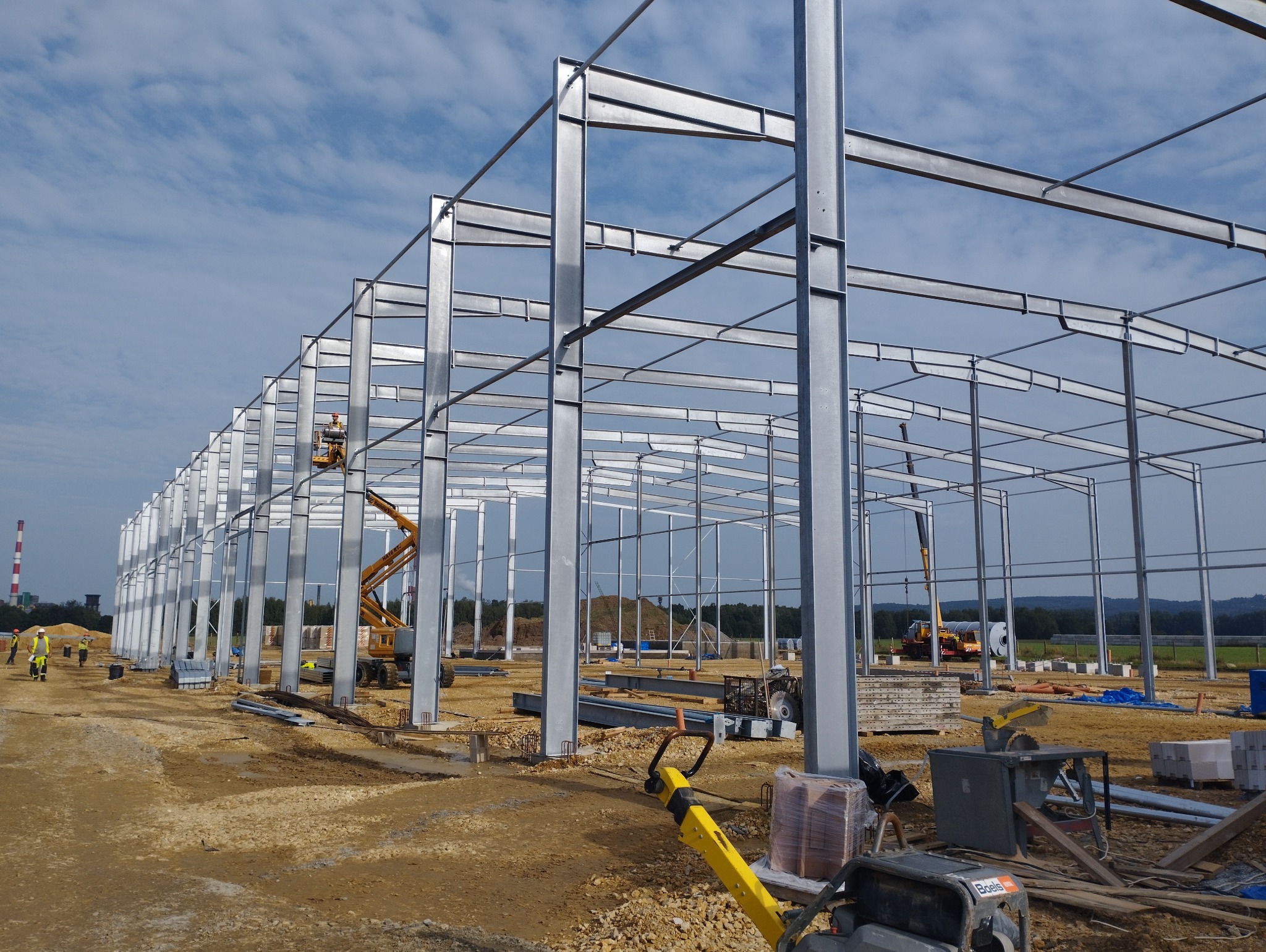 Stalowa konstrukcja hali w trakcie montażu, widoczny dźwig koszowy i pracownicy w oddali, teren budowy z widocznym kominem w tle.
