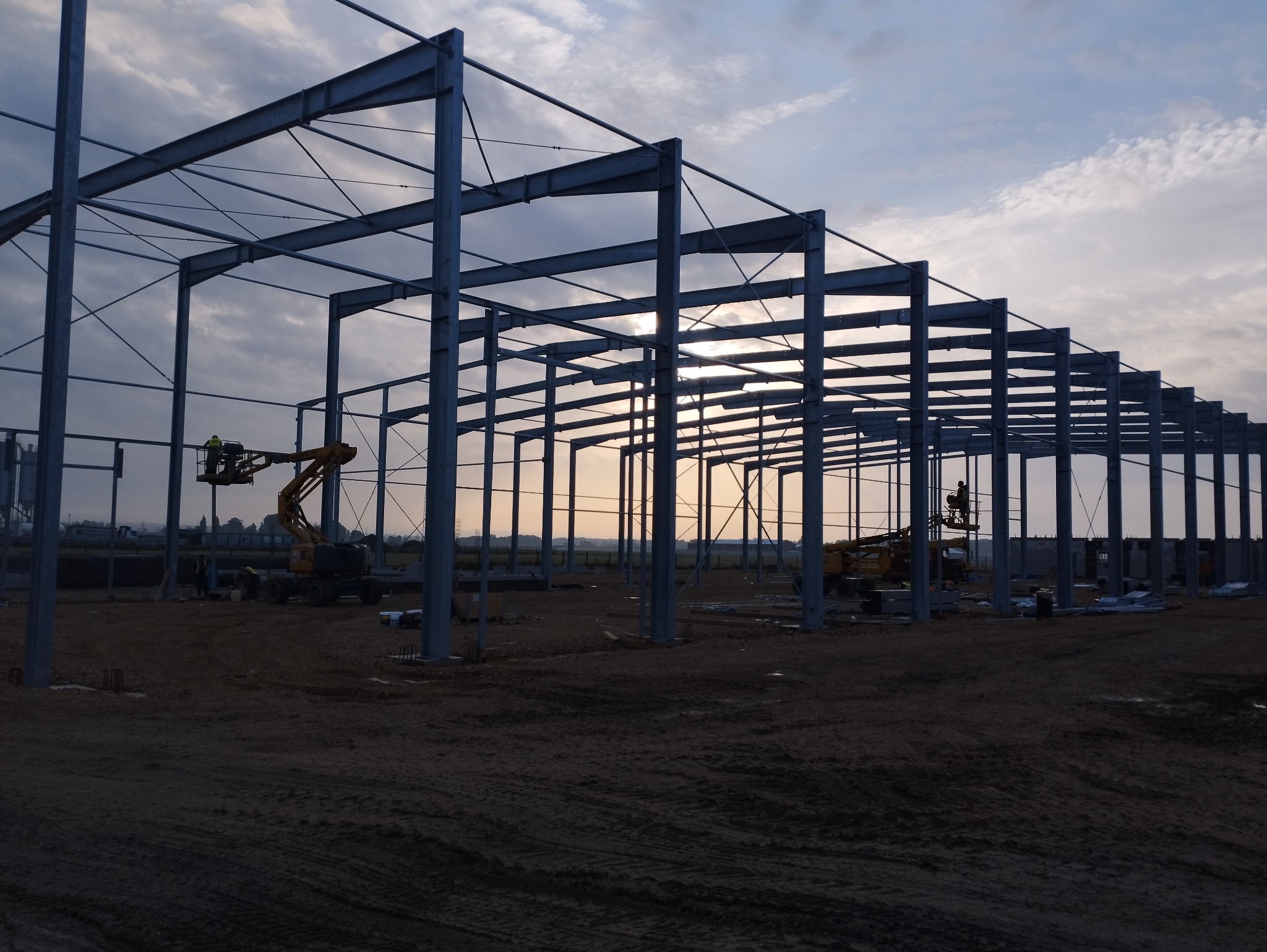 Konstrukcja stalowa hali w trakcie budowy, widoczne słupy i belki, dwie maszyny budowlane z pracownikami na podnośnikach, ziemisty teren budowy, zachmurzone niebo w tle.