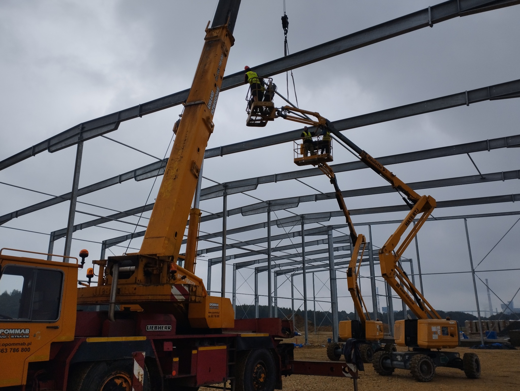 Montaż stalowej konstrukcji hali, widoczne dwie pomarańczowe platformy podnośnikowe i dźwig Liebherr, pracownicy w kamizelkach odblaskowych na wysokościach, pochmurne niebo w tle.