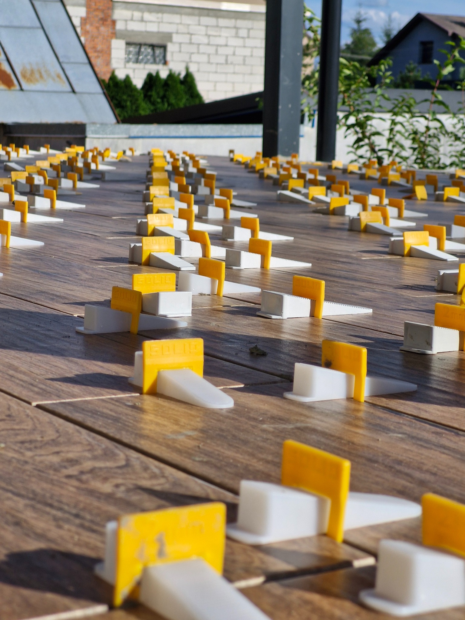 Równomiernie rozmieszczone, żółto-białe kliny poziomujące 'SOLID' na drewnianym tarasie podczas montażu, z widocznymi budynkami w tle.