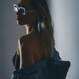 Portret kobiety z profilu, w kwadratowych okularach, z dżinsową kurtką na ramionach i perłowym naszyjnikiem, oświetlona z boku.