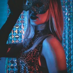 Joanna Janczewska - Kobieta w masce kota z cekinami, pozująca na tle błyszczącej ściany w neonowym oświetleniu.