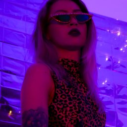 Joanna Janczewska - Kobieta w okularach z kryształkami, w bluzie w panterkę, pozująca na tle ściany z metalowych płytek w fioletowym oświetleniu.