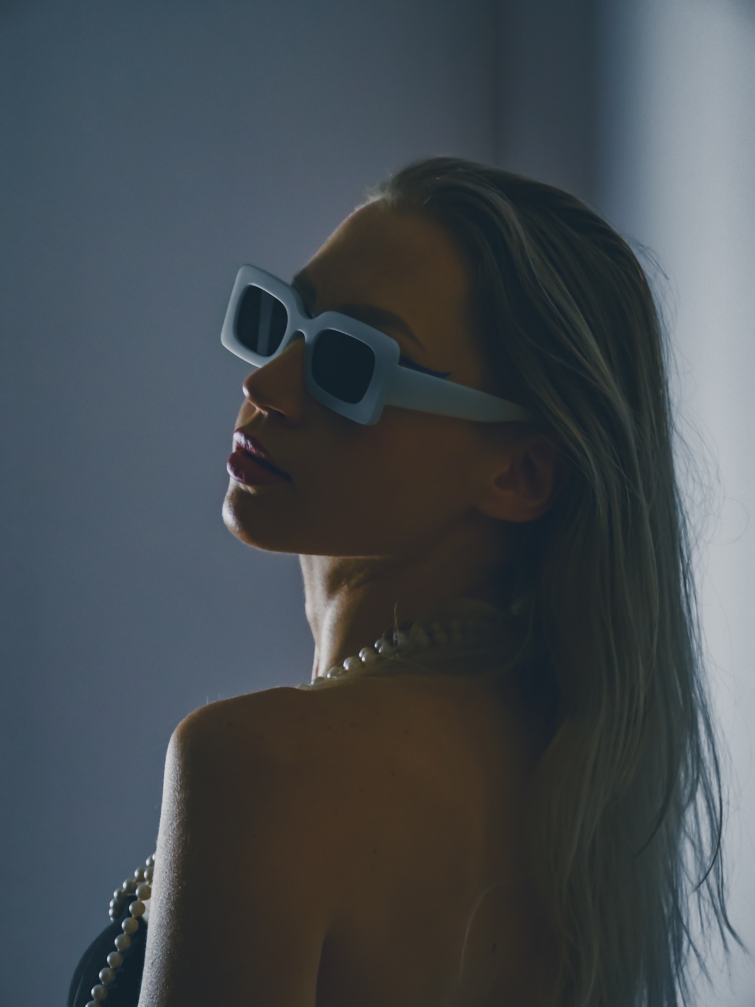 Portret kobiety z profilu, w białych okularach i perłach, z podniesioną głową, na rozmytym tle w chłodnych odcieniach.