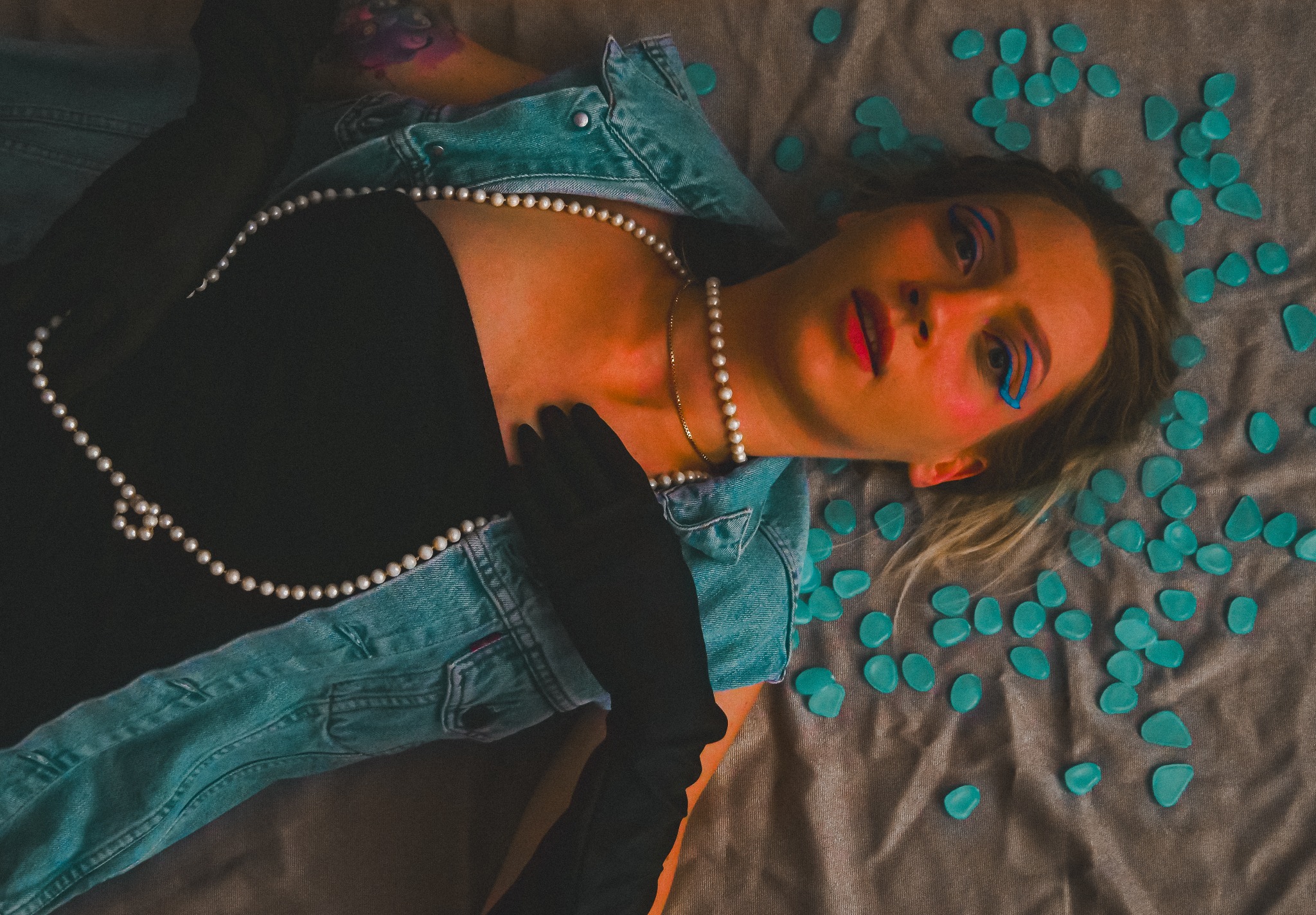 Kobieta leżąca na tkaninie, w dżinsowej kurtce, czarnym topie i rękawiczkach, z perłowym naszyjnikiem, z niebieskim makijażem oczu i koralową szminką, otoczona turkusowymi kamieniami dekoracyjnymi.