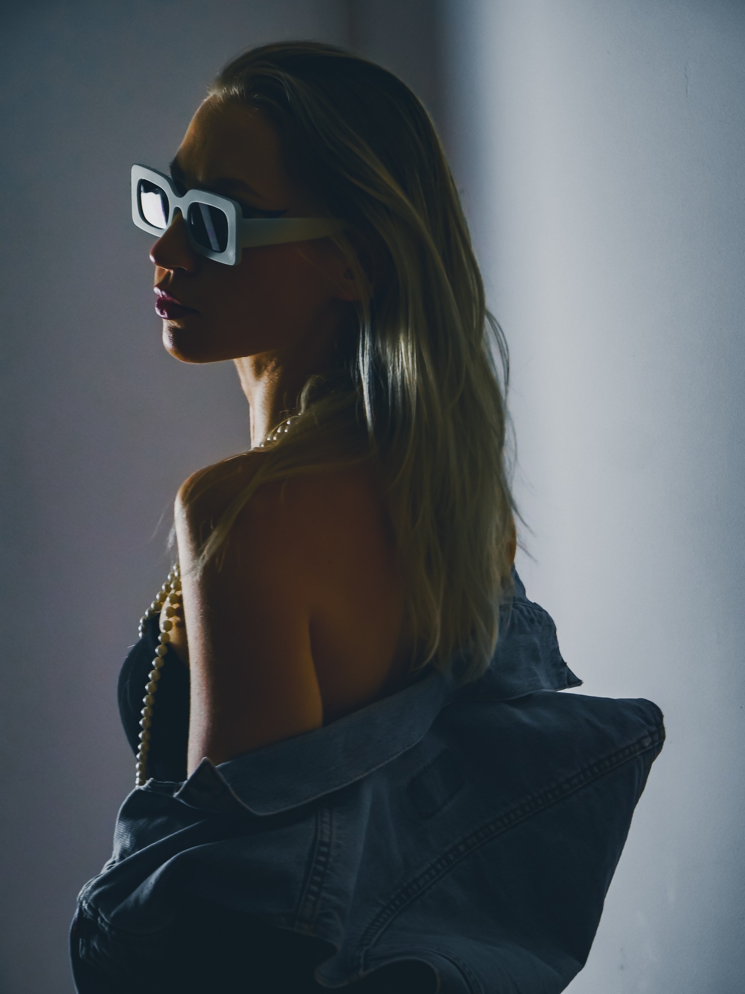 Portret kobiety z profilu, w kwadratowych okularach, z dżinsową kurtką na ramionach i perłowym naszyjnikiem, oświetlona z boku.