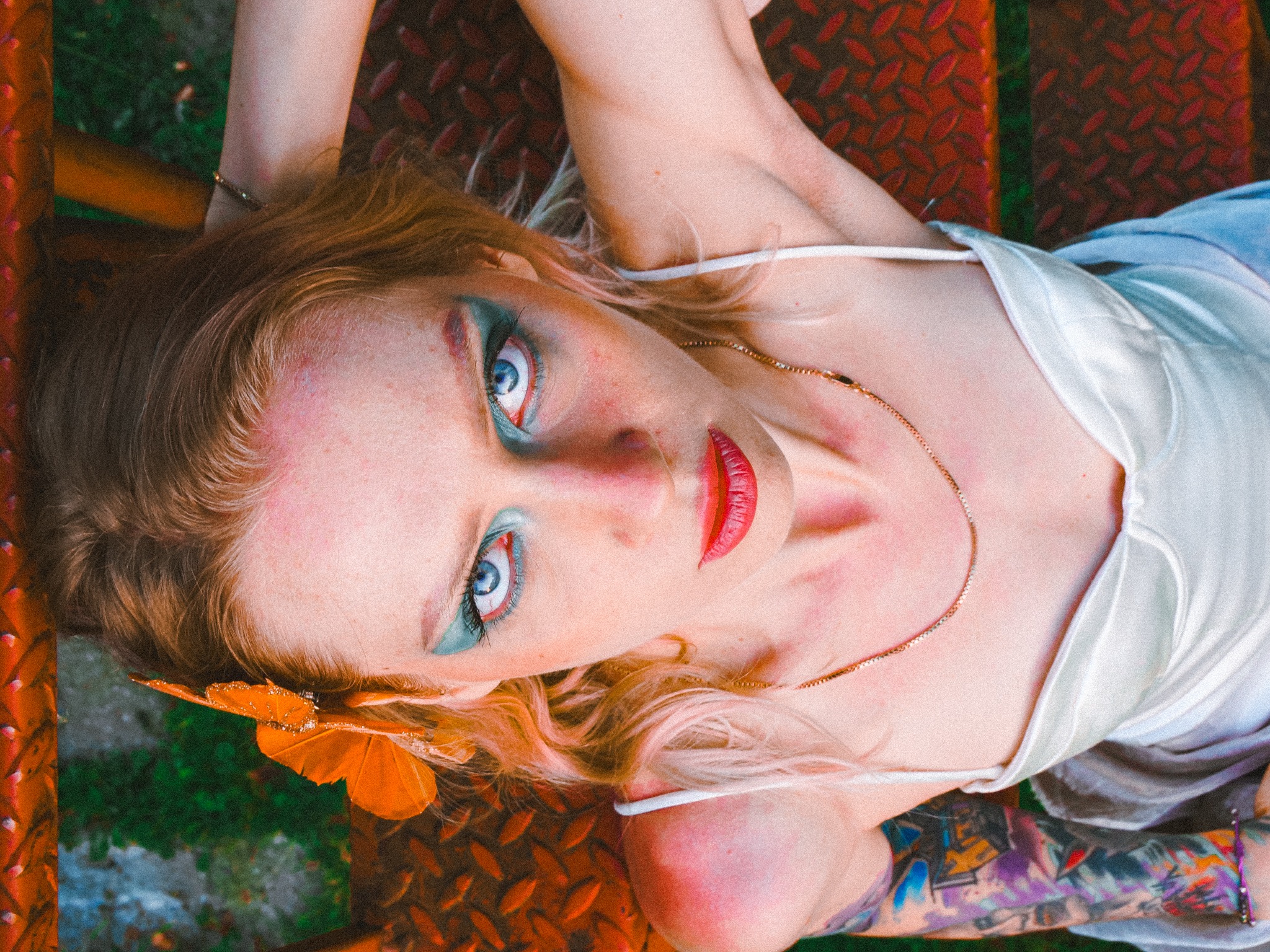 Kobieta z intensywnym makijażem leży na metalowych schodach, widoczne tatuaże na ramieniu, pomarańczowa ozdoba we włosach i niebieskie oczy.