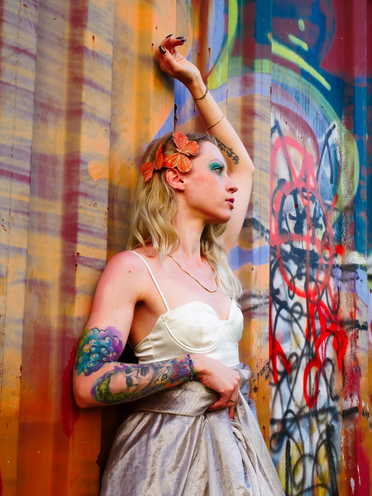 Kobieta w sukience i z makijażem artystycznym pozuje na tle ściany pokrytej graffiti i rdzawymi plamami. Włosy blond, upięte pomarańczowymi motylami. Na ramieniu widoczny tatuaż.