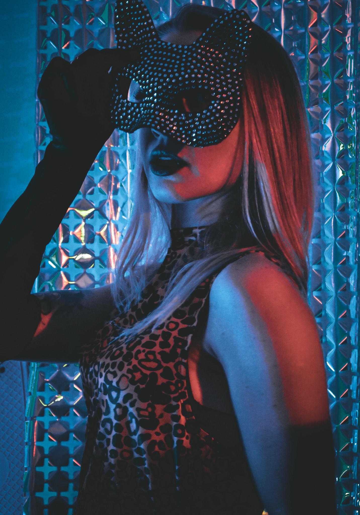Kobieta w masce kota z cekinami, pozująca na tle błyszczącej ściany w neonowym oświetleniu.