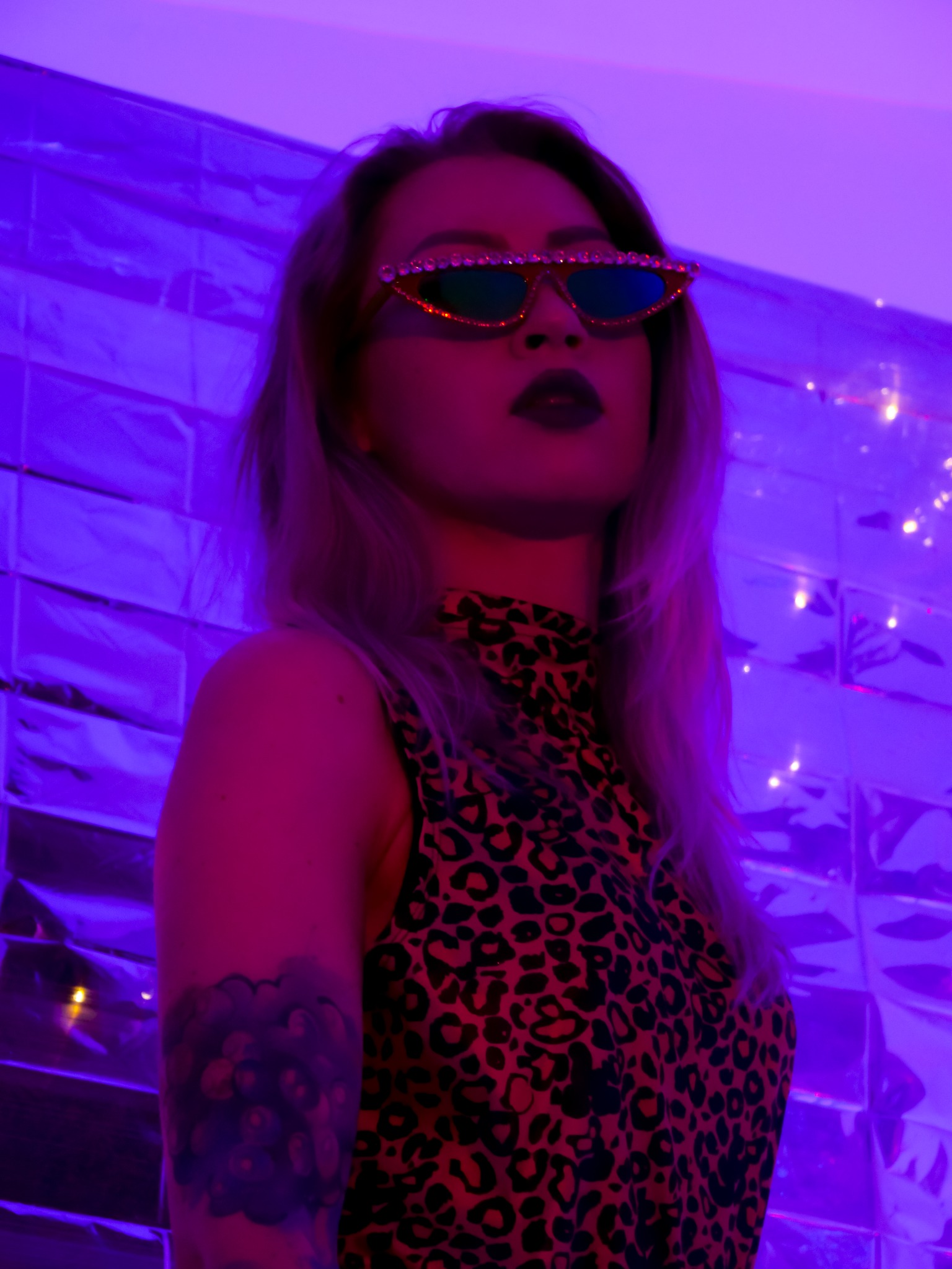 Kobieta w okularach z kryształkami, w bluzie w panterkę, pozująca na tle ściany z metalowych płytek w fioletowym oświetleniu.
