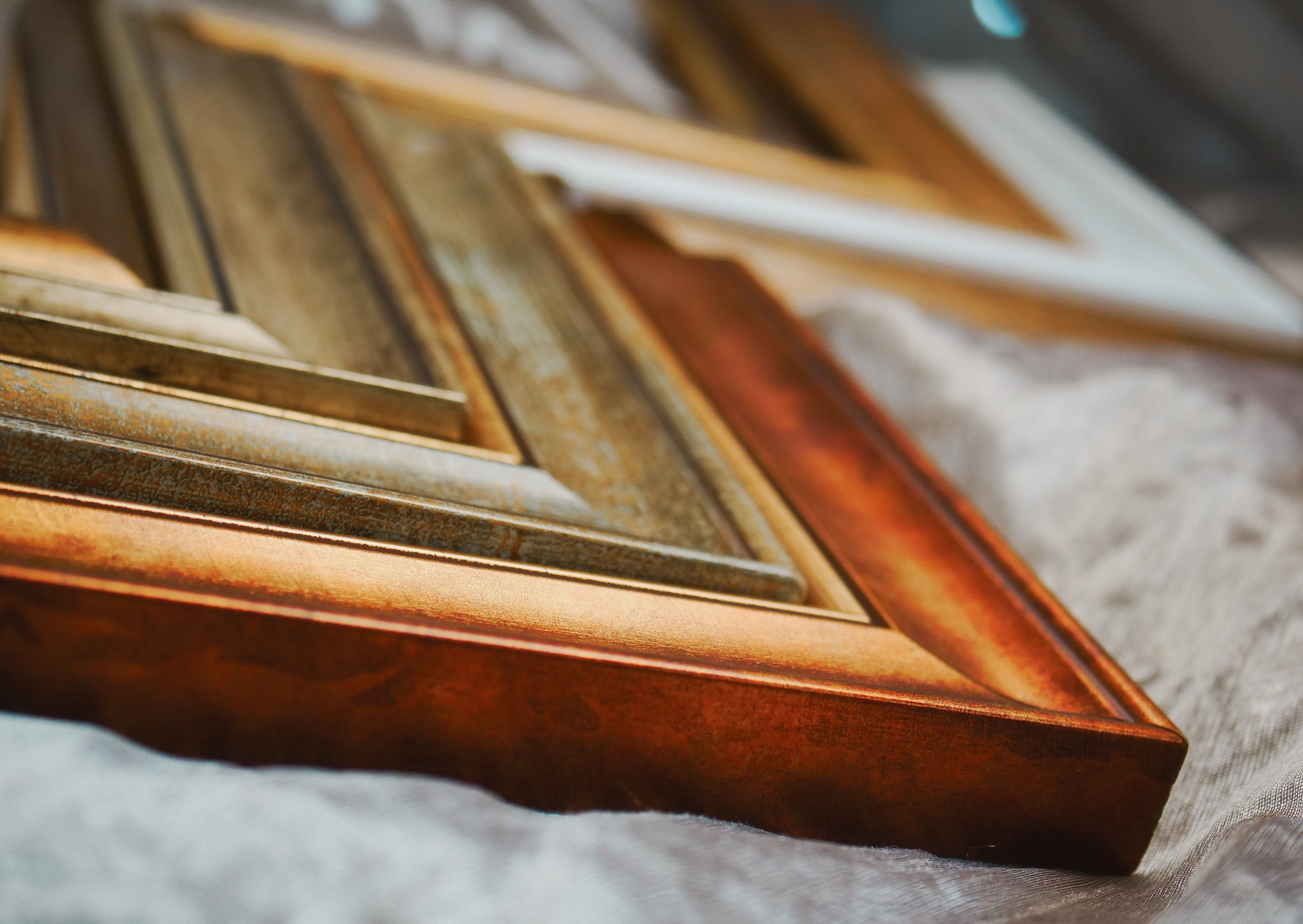 Przekrój przez narożnik ramki do obrazów z Koszalina, ukazujący warstwy i faktury złotej, srebrnej i białej farby na drewnie, leżący na delikatnej, teksturowanej tkaninie.