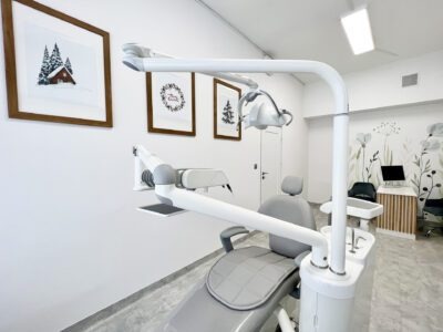 Wnętrze gabinetu z nowoczesnym fotelem dentystycznym w odcieniach szarości, na ścianach grafiki w drewnianych ramkach o tematyce zimowej, białe ściany i minimalistyczny wystrój.