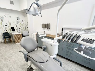 Wnętrze gabinetu z nowoczesnym fotelem dentystycznym w odcieniach szarości, na ścianie dekoracja z motywem roślinnym, w tle biurko z komputerem.