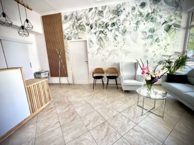 Jasne wnętrze poczekalni kliniki medycznej z recepcją, tapetą w liście, kwiatami na stoliku i miejscami do siedzenia.
