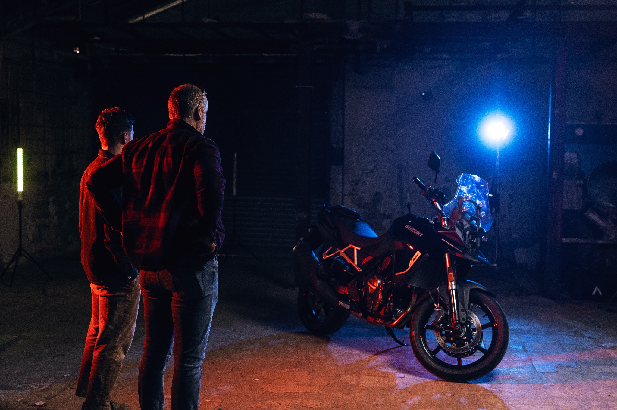 Dwóch mężczyzn obserwuje motocykl Suzuki V-Strom 800DE w studiu z oświetleniem LED i betonową podłogą.