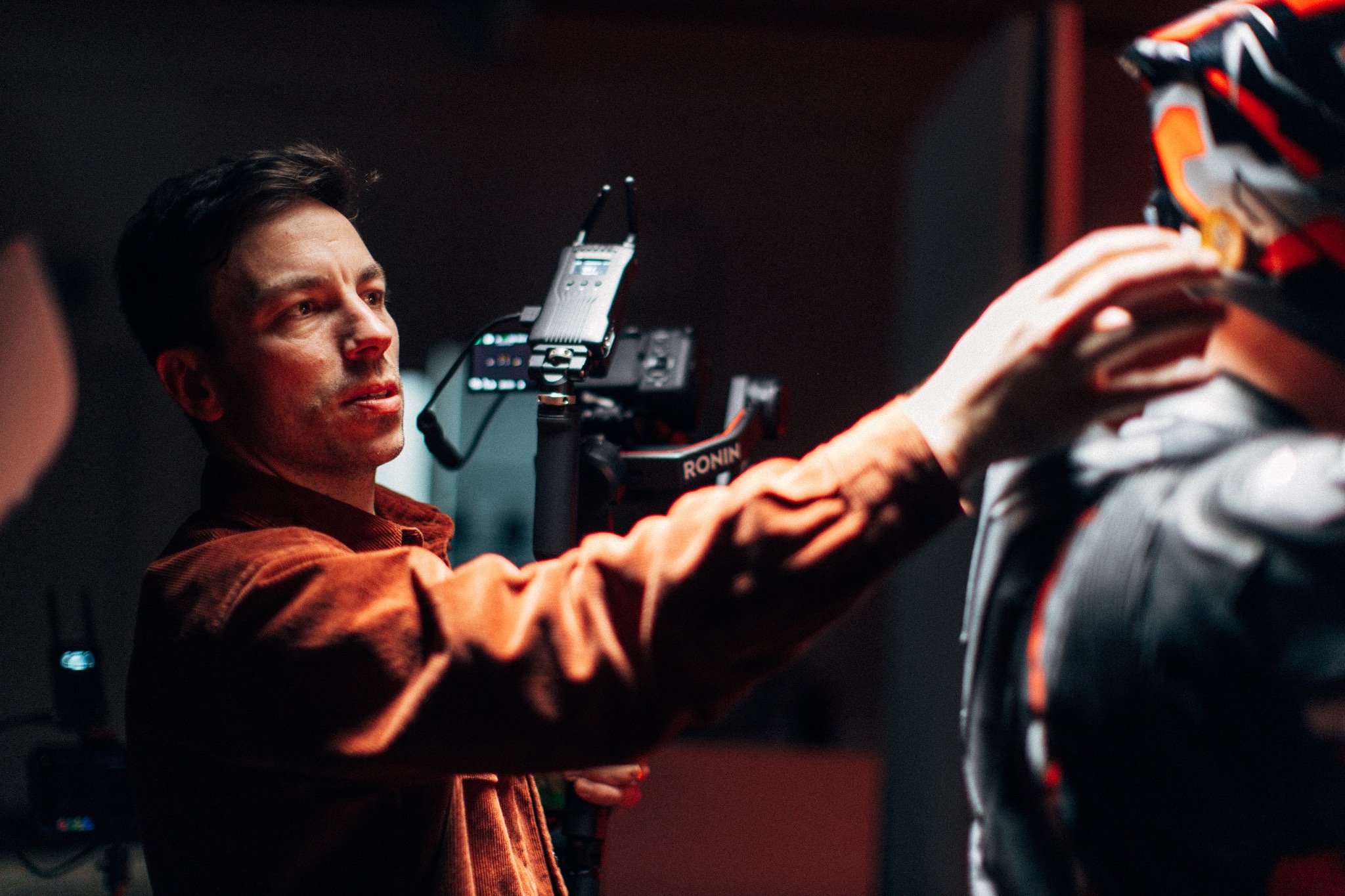 Operator filmowy w brązowej kurtce ustawia mikrofon przy kostiumie postaci podczas kręcenia sceny, widoczny stabilizator kamery Ronin.