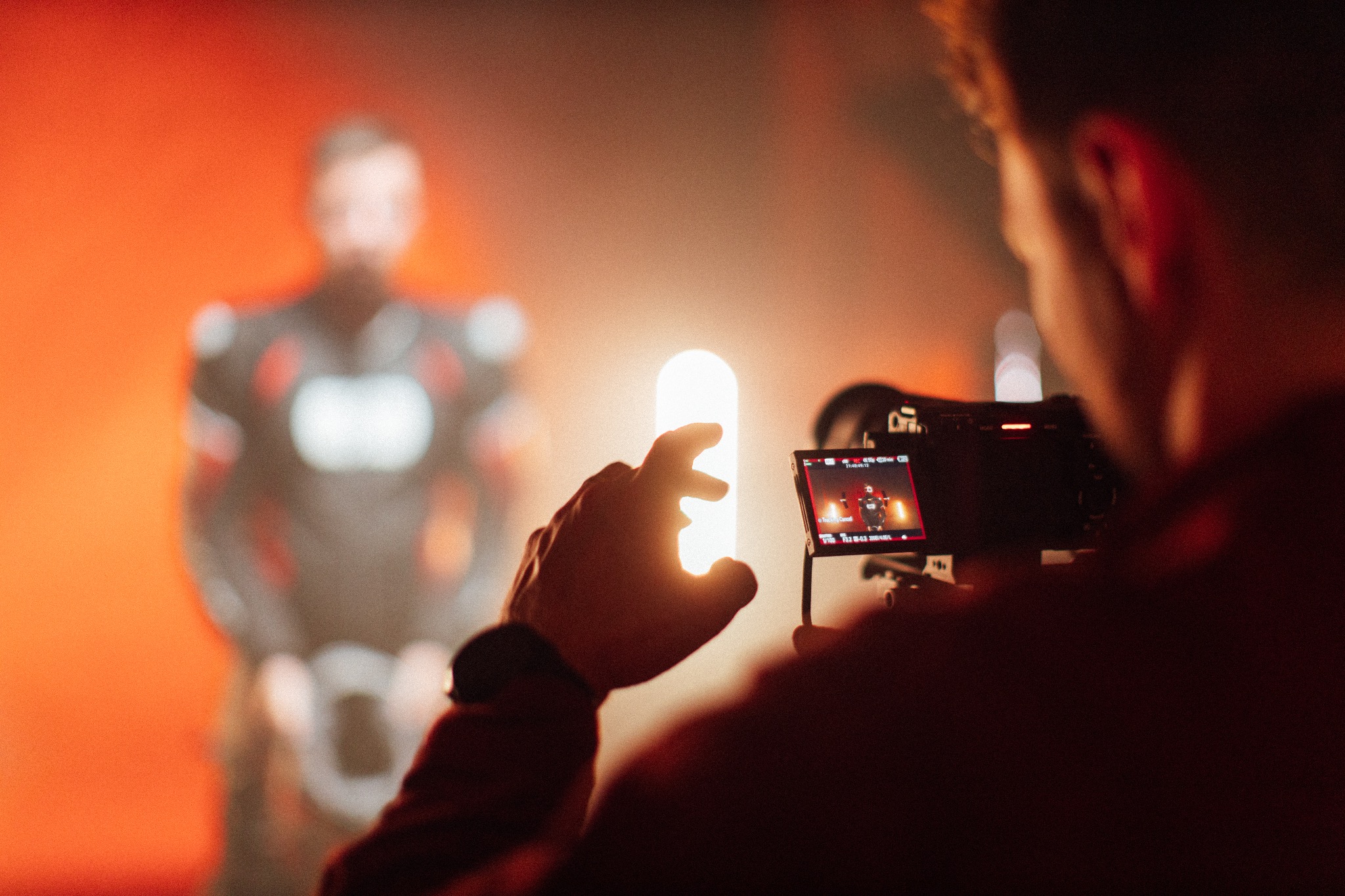 Operator kamery w Oxfordzie filmuje postać w kostiumie na pomarańczowym tle, z użyciem światła LED, widoczne ustawienia ekspozycji na ekranie kamery.