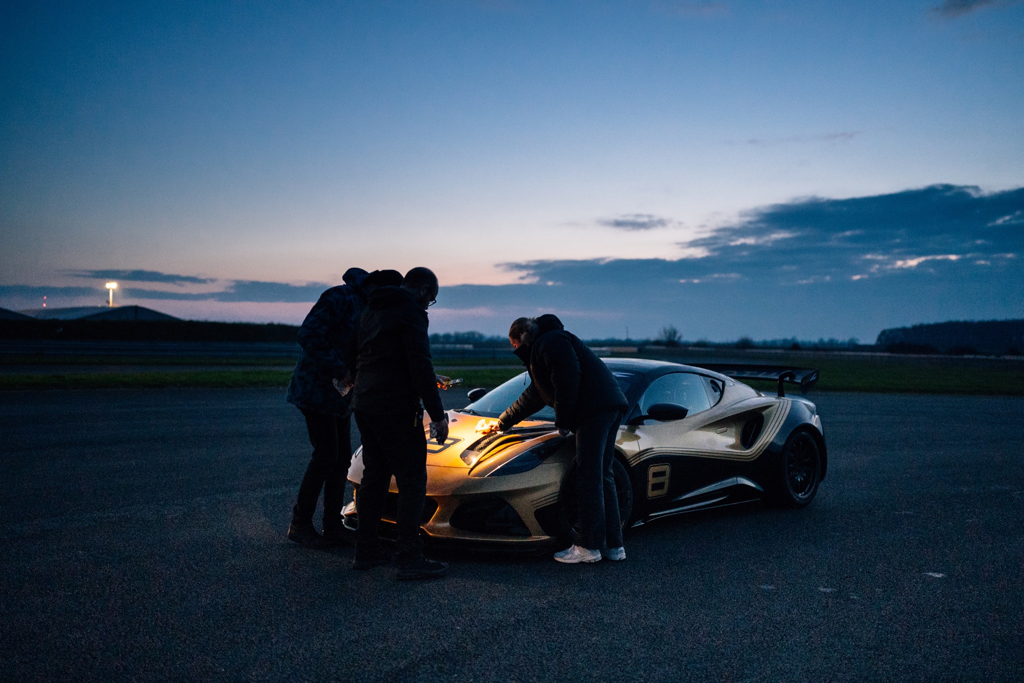 Czterech mężczyzn w ciemnych ubraniach ogląda złoto-czarny samochód sportowy z numerem 8 na boku, stojący na asfalcie o zmierzchu.