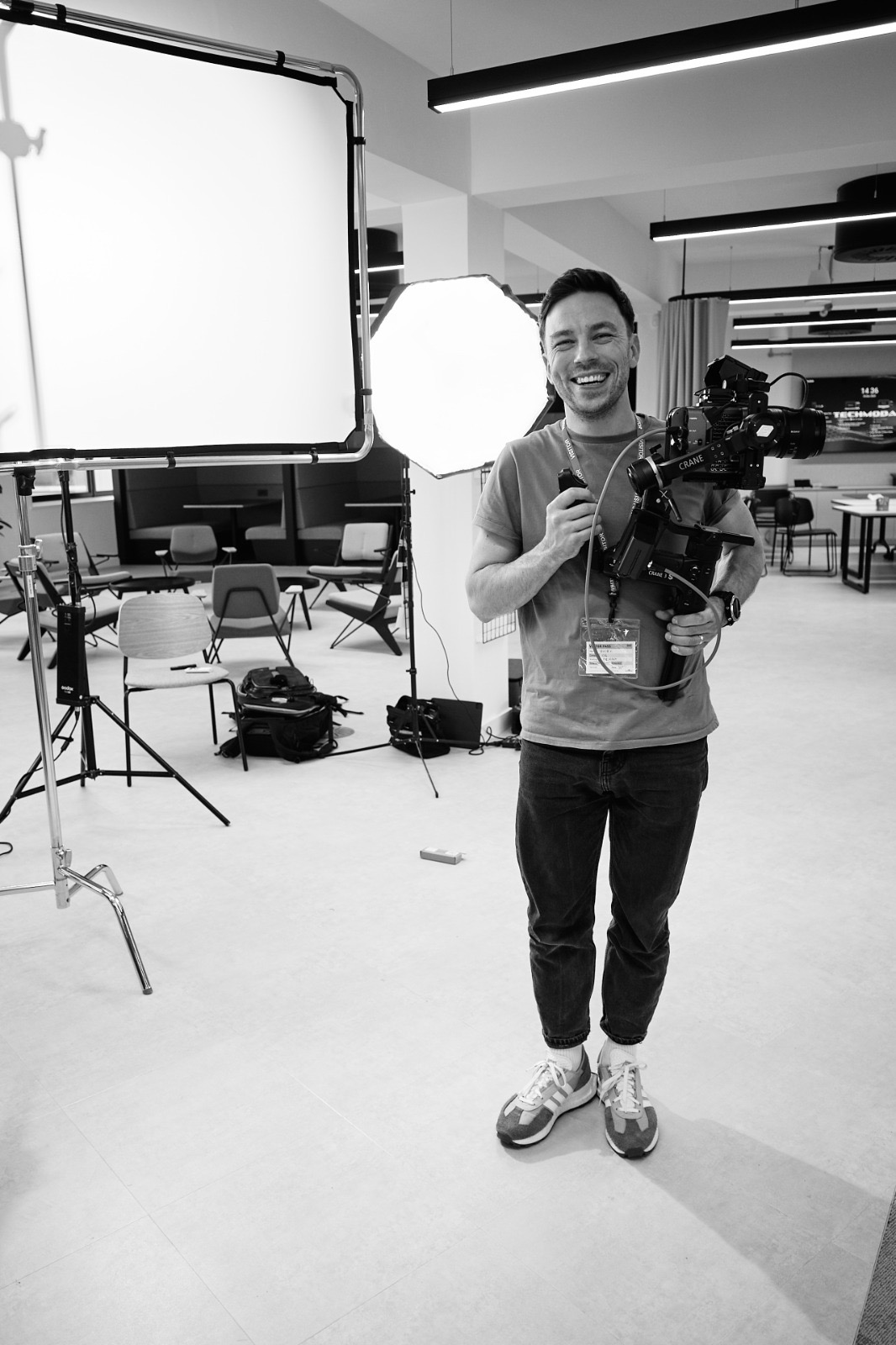 Uśmiechnięty operator kamery z profesjonalnym sprzętem, w tle oświetlenie studyjne i biurowe wnętrze z krzesłami i stołami.