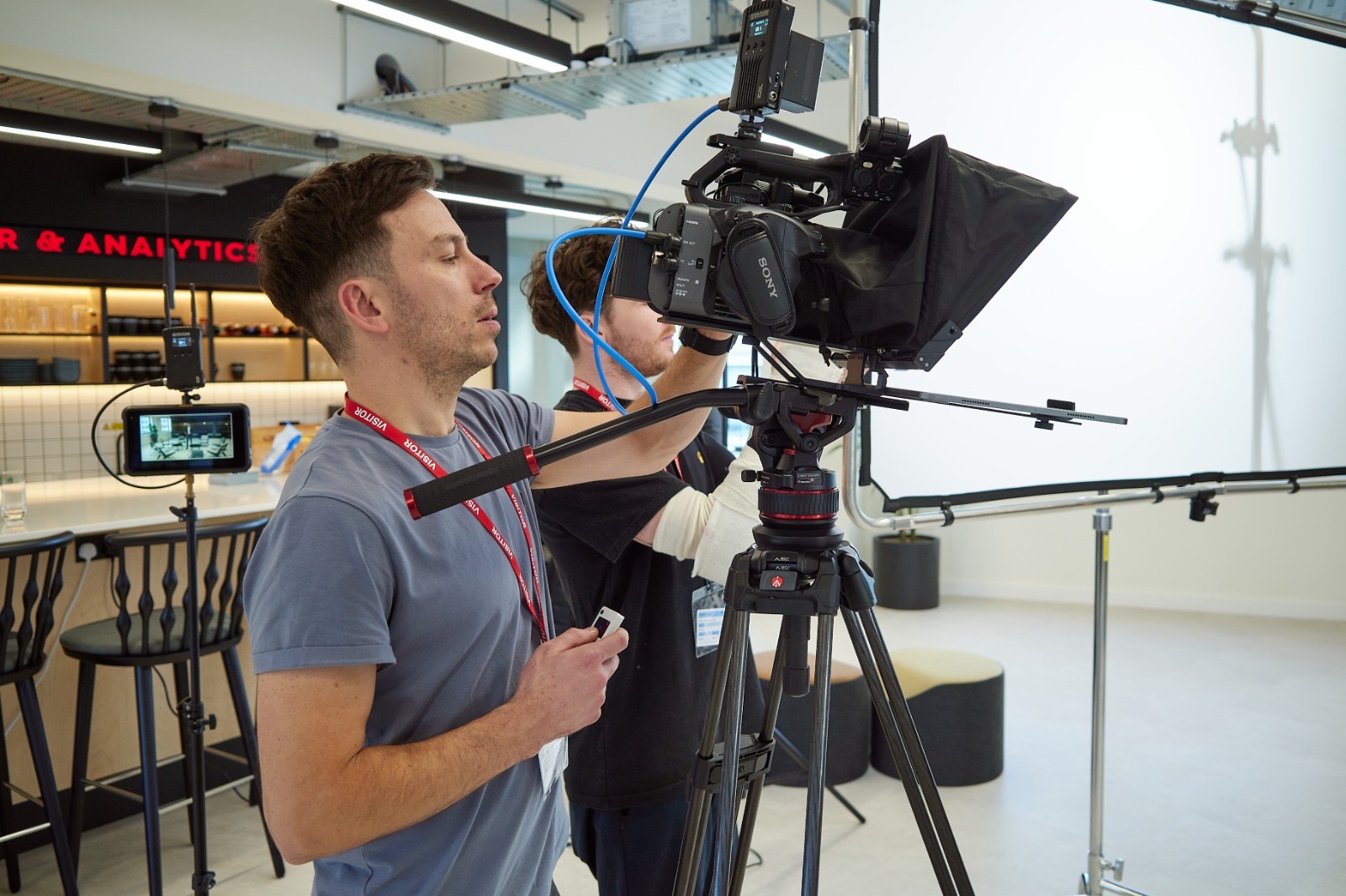 Ekipa filmowa przygotowuje się do nagrania w studio; widoczny operator kamery z identyfikatorem VISITOR na smyczy, profesjonalny sprzęt oświetleniowy i białe tło.