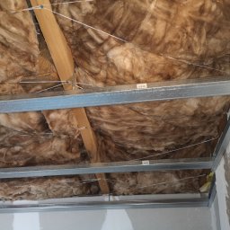 Aircrafttech - Izolacja z wełny mineralnej zamontowana pod sufitem podwieszanym z metalową konstrukcją, widoczne drewniane elementy konstrukcyjne.