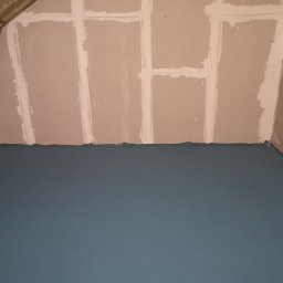 Aircrafttech - Przygotowane do malowania ściany z płyt gipsowo-kartonowych, zabezpieczone taśmą łączenia, z pomalowaną na niebiesko podłogą, widok na narożnik pokoju.