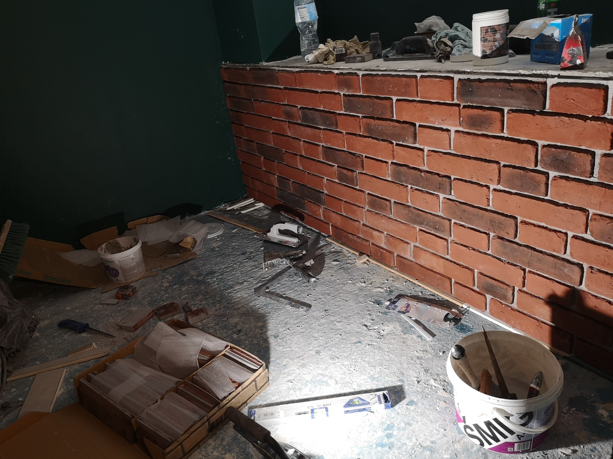 Ściana wykończona cegłą klinkierową w trakcie montażu, widoczne narzędzia, poziomica, opakowania z płytkami, klej i akcesoria budowlane na podłodze.