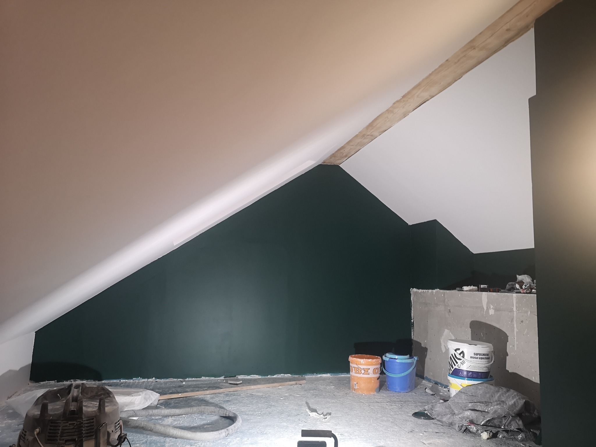 Poddasze po częściowym pomalowaniu ścian na zielono, widoczne wiadro z farbą, odkurzacz i drewniana belka stropowa.