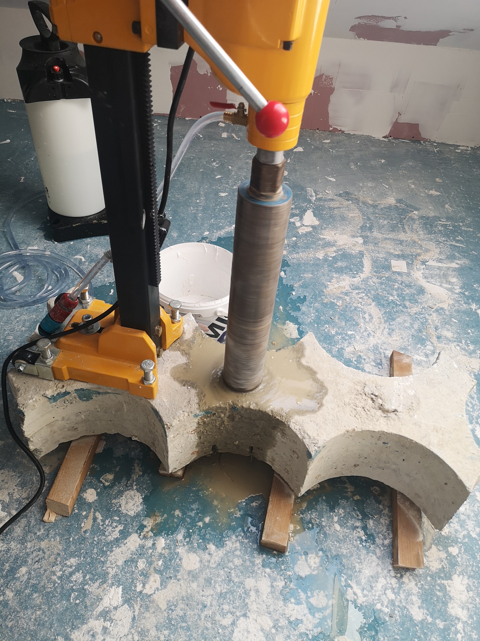 Żółta wiertnica rdzeniowa wiercąca otwór w betonowym elemencie o łukowatym kształcie, podpartym drewnianymi klockami, w pomieszczeniu z pobrudzoną podłogą.